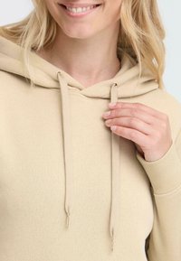 Felpa beige con cappuccio regolabile, polsini a costine e texture liscia, caratterizzata da un design semplice e dettagli minimi.
