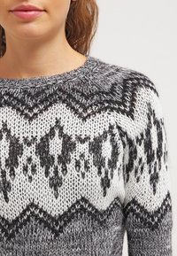 Donna che indossa un maglione lavorato a maglia grigio e bianco con motivi geometrici neri, vista dalle spalle alle labbra su uno sfondo neutro.