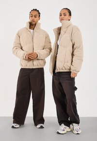 Vestes en duvet en velours côtelé beige avec cols hauts et fermetures éclair, associées à des pantalons larges sombres et des baskets. Tissu texturé et détails d'ourlet élastique.