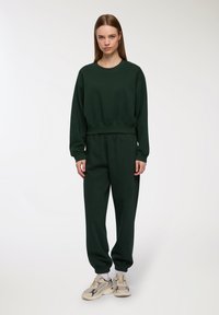 Ciemnozielona, bawełniana bluza z okrągłym dekoltem i elastycznym pasem. W zestawie z dopasowanymi, zwężającymi się dresami i beżowymi sneakersami.