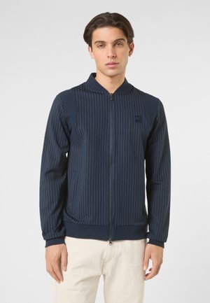 Mężczyzna stojący, ubrany w granatową marynarkę na suwak w paski z prążkowanymi mankietami i kołnierzem, w połączeniu z jasnymi spodniami.