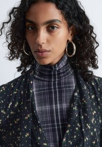 Black floral-patterned fabric with small pink flowers over a dark plaid turtleneck in purple tones; large silver hoop earrings visible.
