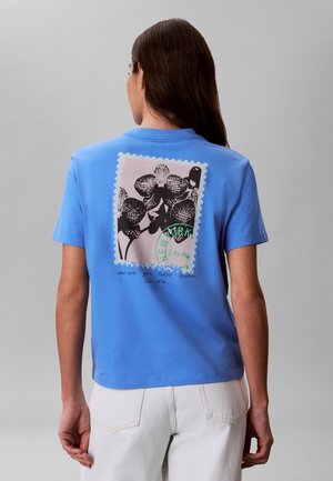 Femeie cu păr lung și castaniu care poartă un tricou albastru cu un imprimeu floral alb-negru și numele orașelor imprimate pe spate, asortat cu pantaloni albi.