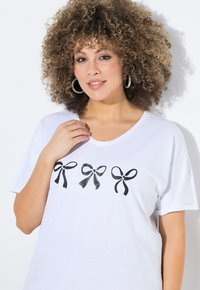 T-shirt blanc avec un col en V présentant trois graphismes de nœuds noirs. Tissu doux, manches courtes et coupe décontractée.