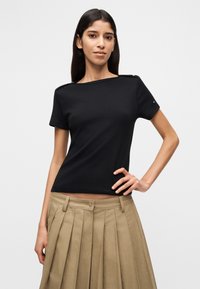 Μαύρο κοντομάνικο τοπ με τετράγωνο ντεκολτέ και κουμπιά στους ώμους. Συνδυάζεται με μπεζ πλισέ φούστα, παρουσιάζοντας ραμμένη εφαρμογή.