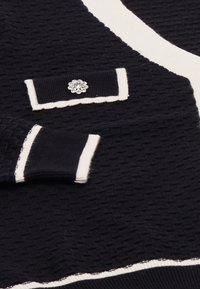 Mustikka tekstuurinen neule paksu valkoinen reunus, jossa on koristeellinen tasku kukkakuvioisella strassikoristeella ja ribatut hihansuut.