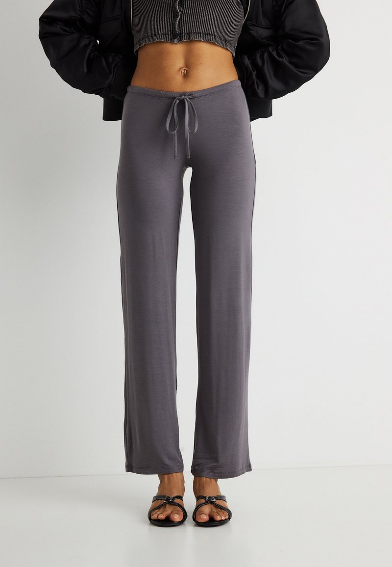 Γυναίκα που φορά γκρι παντελόνι lounge με κορδόνια, μαύρο κοντό τοπ, μαύρο σακάκι και μαύρα σανδάλια με ανοιχτά δάχτυλα, στέκεται μπροστά σε λευκό φόντο.