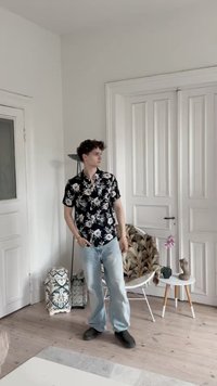 Camicetta nera a fiori a maniche corte, jeans azzurri chiari e scarpe nere, sullo sfondo neutro con una lampada e oggetti decorativi.