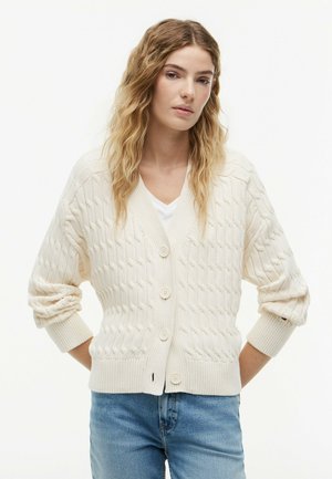 Jeune femme portant un cardigan blanc en maille torsadée boutonné sur une chemise blanche et un jean bleu, debout les mains dans le dos.