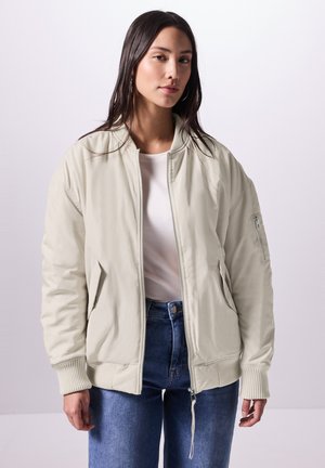 Young woman wearing a light beige bomber jacket over a white shirt and blue jeans, standing against a plain light background.