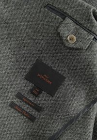 Tissu texturé gris avec un motif en chevrons, comportant un bouton et plusieurs étiquettes pour les informations de taille et de marque.