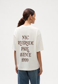 Mujer con pelo castaño en una coleta, que lleva pendientes de aro dorados, una camiseta blanca de gran tamaño con el texto "NYC Riverside Park Avenue 1999" en marrón, y jeans azules.