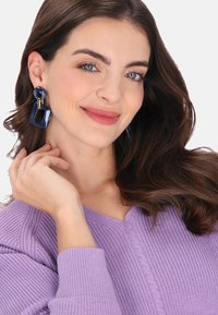 Lila bordázott kötött pulóver V-nyakkivágással. A modell nagy, tengerészkék statement fülbevalót visel geometrikus alakzatokkal és arany díszítéssel.