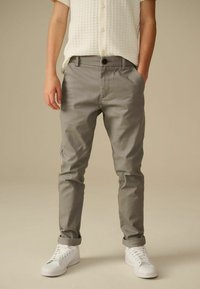 Pantalon en coton gris avec une coupe décontractée, présentant une fermeture à bouton et des ourlets retroussés, associé à des baskets blanches. Chemise blanche à manches courtes texturée.