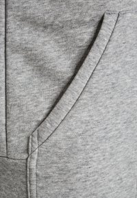 Primo piano di un tessuto di felpa grigia, caratterizzato da una consistenza morbida, un sottile motivo melangiato e una tasca frontale con cucitura a punto.