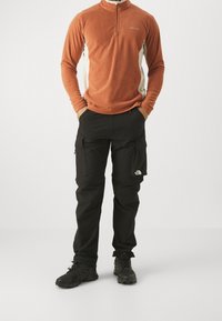 Polarowy sweter z zamkiem ćwierć w kolorze rdzy z kremowymi panelami bocznymi, zestawiony z czarnymi spodniami cargo z kieszeniami i logo. Czarny obuwie outdoorowe dopełnia całość.