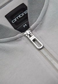 Sudadera gris con cremallera y tejido texturizado. Presenta una cremallera plateada y una etiqueta negra marcada con "ombre" y la talla "M" cerca del cuello.