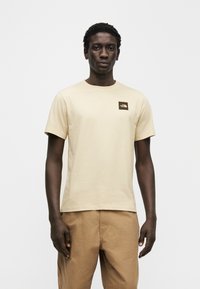 Ανοιχτό μπεζ κοντομάνικο T-shirt από μαλακό βαμβάκι, με σκοτεινό καφέ λογότυπο στο αριστερό στήθος. Απλός, καθημερινός σχεδιασμός.