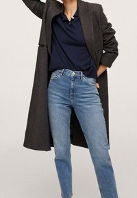 Un lungo cappotto nero a scacchi sopra una polo blu navy, abbinato a jeans a vita alta di un blu chiaro. L'outfit presenta linee semplici e tagli classici.