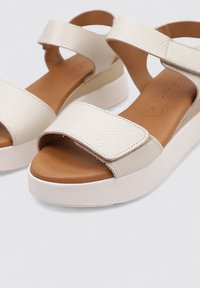 Sandalias off-white hechas de un material sintético texturizado con una plantilla marrón, que cuentan con una tira ancha y una suela de cuña.