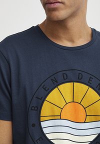 Marineblå bomulds T-shirt med et cirkulært grafisk design, der har en gul sol og bølgede linjer i hvid og blå, sort omrids med tekst.