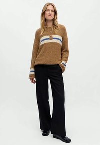 Žena oblečená v hnědém chlupatém svetru s bílými a modrými pruhy, černých širokých kalhotách a černých botách, stojící s jednou rukou v kapse.