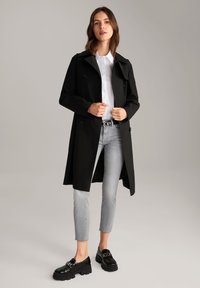 Fekete hosszú trench coat, dupla gombolású dizájnnal, fehér inggel és szürke farmernadrággal párosítva. Fényes fekete cipő jellemzi, vastag talppal.