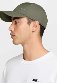 Olíva zöld baseball sapka, amely szövetből készült, ívelt napellenzővel és ezüst Nike logóval. A modellen fehér póló van, amelyen egy kicsi fekete logó látható.