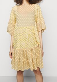 Żółta sukienka w wzory z krótkimi, rozkloszowanymi rękawami i warstwowym, falbaniastym dołem. Materiał zdobiony jest geometrycznymi kształtami w subtelnych odcieniach.