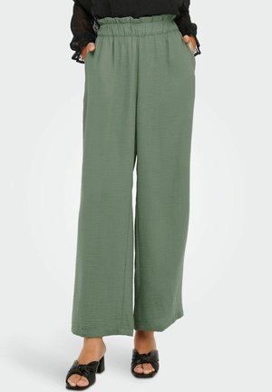 Femme portant un pantalon vert olive taille haute, ample, avec des poches latérales, et des sandales à talons noires à bouts ouverts, debout les mains dans les poches.