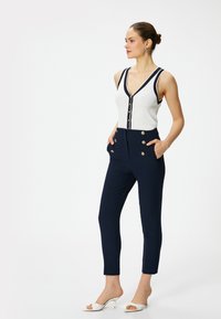 Marineblauwe high-waisted broek met goudkleurige knopen, gecombineerd met een witte geribde tanktop met een diepe V-hals en contrasterende bies.
