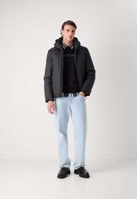 Svart pufferjakke med hette, glidelås, kombinert med svart genser med rund hals og lyseblå jeans, avsluttet med svarte sko.
