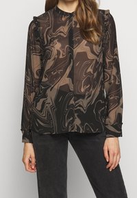 Vrouw in een doorzichtige blouse met lange mouwen, voorzien van bruin en zwarte spiralen marmerpatronen en geraffineerde schouderdetails, gecombineerd met zwarte broek.