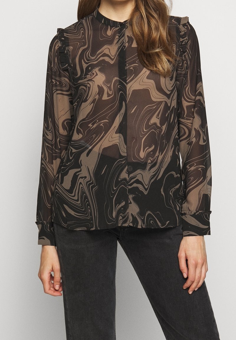 Vrouw in een doorzichtige blouse met lange mouwen, voorzien van bruin en zwarte spiralen marmerpatronen en geraffineerde schouderdetails, gecombineerd met zwarte broek.