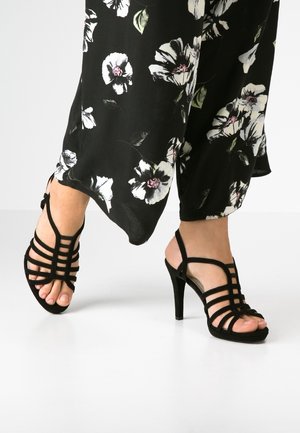 Pieds portant des sandales noires à talons hauts avec des brides, associées à un pantalon noir avec des motifs floraux blancs et roses.