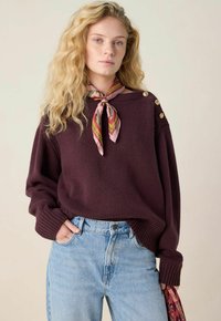 Junge Frau mit langen, lockigen blonden Haaren, die einen kastanienbraunen Pullover mit goldenen Knöpfen, einen pinken, gemusterten Halstuch und eine hellblaue Jeans trägt.