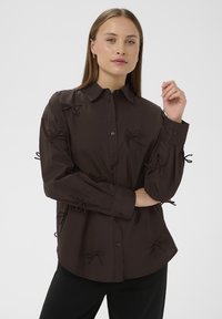 Brun skjorte med knapper og sløjfer på ærmerne, lavet af letvægtsstof, med en buet kant og lange ærmer.
