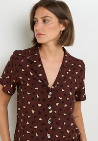 Chemise à manches courtes, boutonnée, de couleur marron avec un motif léopard en beige et noir. Dispose d'un col classique et d'un tissu léger et lisse.