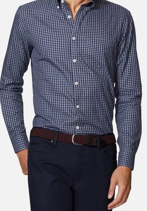 Mężczyzna w niebiesko-białej koszuli w kratę zapiętej na guziki, wsuniętej w ciemne spodnie z czarno-czerwonym tkanym paskiem.