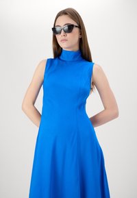 Blaues ärmelloses Kleid mit hohem Kragen, das über eine glatte, weiche Textur und eine A-Linie-Silhouette verfügt. Begleitet von schwarzen, kantigen Sonnenbrillen.