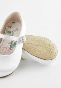 Ballerines blanches avec une sangle, ornées d'une boucle en cristal. Dotées d'une semelle intérieure florale et d'une semelle en caoutchouc texturée et à motifs.