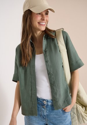 Femme souriante portant une casquette beige, une chemise verte à manches courtes boutonnée sur un haut blanc, un jean bleu, et portant un sac fourre-tout beige sur l'épaule.