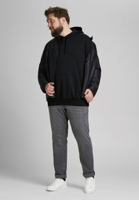 Must langade musta kapuutsiga sweatshirt musta jope all, koos keeratud hallide teksade ja valgete spordijalatsitega. Pehmed materjalid ja lõdva istuvusega.