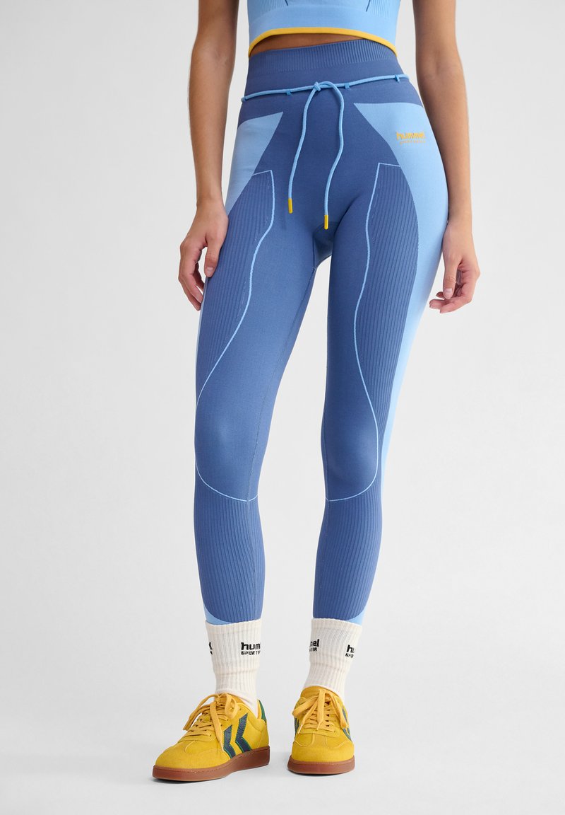 Blå och ljusblå leggings med ribbad textur, med dragsko i midjan och geometrisk paneldesign, tillsammans med gula sneakers.
