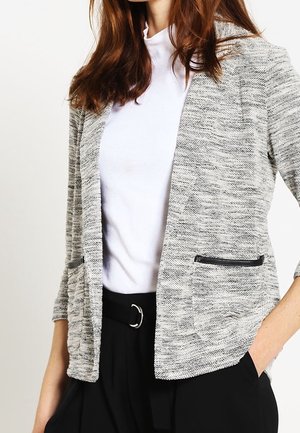 Vrouw draagt een gestructureerde grijze open blazer met zwarte ritszakken, witte geribbelde coltrui en zwarte broek met riem, hand in de zak.