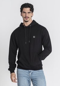 Felpa nera con cappuccio realizzata in tessuto morbido. Presenta una tasca frontale, cappuccio con cordino e un piccolo logo sulla parte sinistra del petto. Abbinata a jeans chiari.