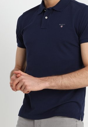 Mann trägt ein marineblaues GANT-Poloshirt mit kurzen Ärmeln, steht mit verschränkten Händen, hellgraue Hose sichtbar.
