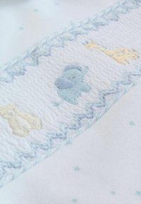 Tessuto bianco con motivi animali ricamati in tonalità pastello di blu e giallo: un elefante, una giraffa e un orso, separati da un bordo sagomato a zigzag.