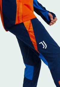Trainingsanzug mit einem blauen und orangefarbenen Design, aus glattem Stoff mit geometrischen Mustern gefertigt. Beinhaltet ein weißes Logo am Bein.