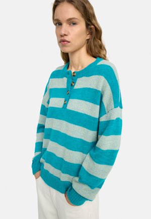 Kobieta o falowanych włosach, ubrana w szeroki sweter w turkusowo-szare paski z drewnianymi guzikami, ręce w kieszeniach białych spodni, neutralne tło.